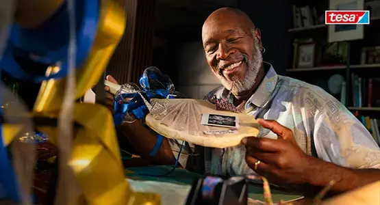 Ein lächelnder älterer Mann, der ein gerahmtes Kunstwerk an einem Basteltisch präsentiert, umgeben von tesa Klebeprodukten.