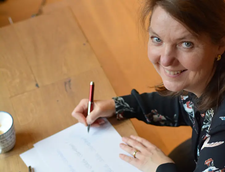 Eine Frau, die auf ein Blatt Papier schreibt, lächelt in die Kamera.