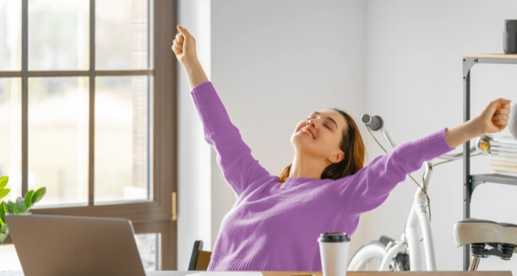 Das Bild zeigt eine Frau in einem Büro, die sich entspannt und lächelnd zurücklehnt, während sie beide Arme nach oben streckt. Sie trägt einen lilafarbenen Pullover und sitzt an einem Schreibtisch mit einem Laptop, Büchern, Notizblock und einer Tasse Kaffee. Im Hintergrund sind ein großes Fenster, ein Regal und ein Fahrrad zu sehen. Der Fokus liegt auf dem Thema Gesundheit und Entspannung im Büro.
