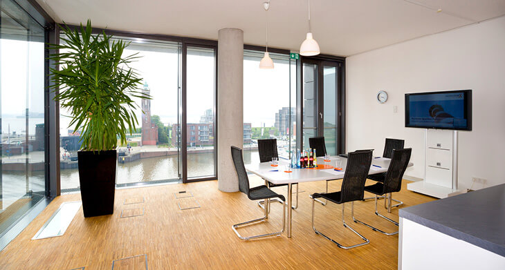Blick in einen Konferenzraum mit bodentiefen Fenstern. Draußen sieht man einen breiten Wasserlauf, innen steht links eine große Topfpflanze, rechts ein Konferenztisch mit 6 Stühlen und einem Monitor an der Wand. Helles Parkett ist auf dem Boden verlegt.