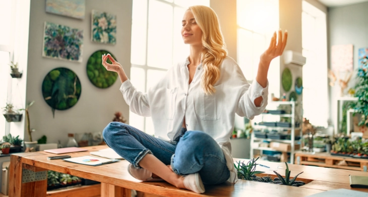 Frau sitzt entspannt im Schneidersitz auf einem Schreibtisch und meditiert.