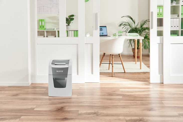 Das Bild zeigt einen Leitz Aktenvernichter in einem modernen Büro, der zentral vor einem offenen Büroraum steht. Im Hintergrund ist ein Schreibtisch mit Bürozubehör sowie ein Stuhl zu sehen.