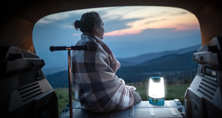 Frau sitzt im Van mit Decke und Campingleuchte beim Sonnenuntergang.
