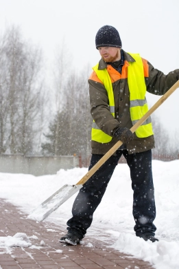 Arbeiter in Warnweste und Mütze räumt Schnee mit einer Schneeschaufel auf einem Gehweg..