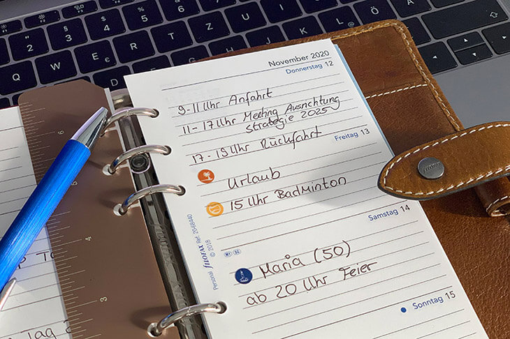 Ein aufgeschlagener Kalender von Filofax liegt auf einem Tisch, im Hintergrund sieht man eine Tastatur. Auf dem Kalenderblatt sieht man verschiedene Einträge und kleine Sticker mit Icons, ein Kugelschreiber ragt ins Bild.  