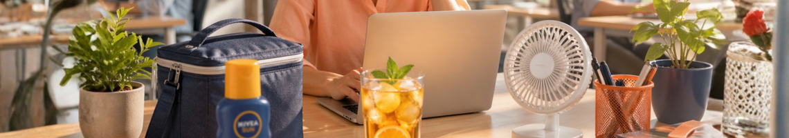 Eine Frau sitzt am Tisch mit Kühltasche, Glas, Ventilator und Sonnencreme.
