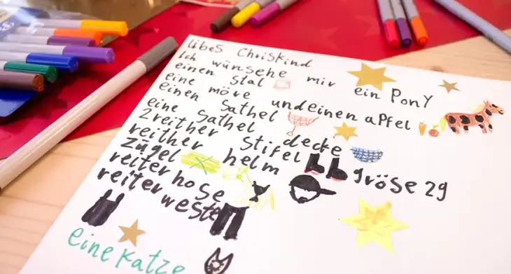 Auf einem Tisch liegen ein von einem Kind geschriebener und gemalter Wunschzettel an das Christkind. Daneben liegen viele Buntstifte.