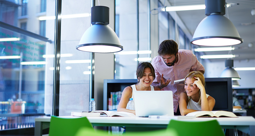 Zwie Frauen und ein Mann sind in einem Großraum-Büro und schauen zusammen auf einen Laptop und unterhalten sich.