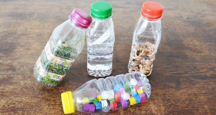 4 kleine Plastikflaschen mit rotem, grünem, gelbem und lila Deckel sind mit Wasser, Gras, Nuss-Schalen und kleinen Holzklötzchen gefüllt.