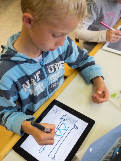 Ein Junge im blau-gestreiften Pullover sitzt an einem Tisch. Mit Hilfe eines Stylus von Online malt er auf einem Tablet ein Bild von einem Krokodil. Im rechten Bildrand sieht man ein weiteres Kind mit einem Tablet sitzen.