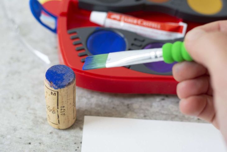 Eine Hand bestreicht Weinkorken mit blauer Farbe vor rotem Aquarellkasten