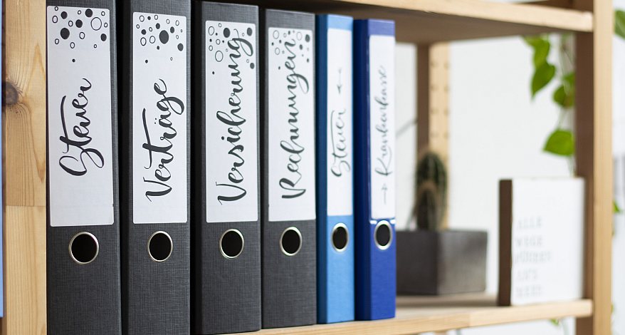 In einem Regal aus hellem Holz stehen einige schwarze und blaue Ordner mit verschiedenen Breiten. Diese wurden mit Ordnerrückenetiketten von Avery Zweckform beschriftet. Darauf zu lesen sind geletterte Begriffe wie "Steuer", "Verträge" oder "Versicherung". Am rechten Bildrand sind unscharf einige Pflanzen zu erkennen.