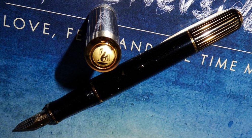 Das Füllermodell Ductus der Marke Pelikan liegt auf einem blauen Untergrund. Der Füller ist schwarz und hat einige goldene Verzierungen. Die Kappe steht aufrecht und im Deckel ist das Pelikan-Logo in Gold eingraviert.