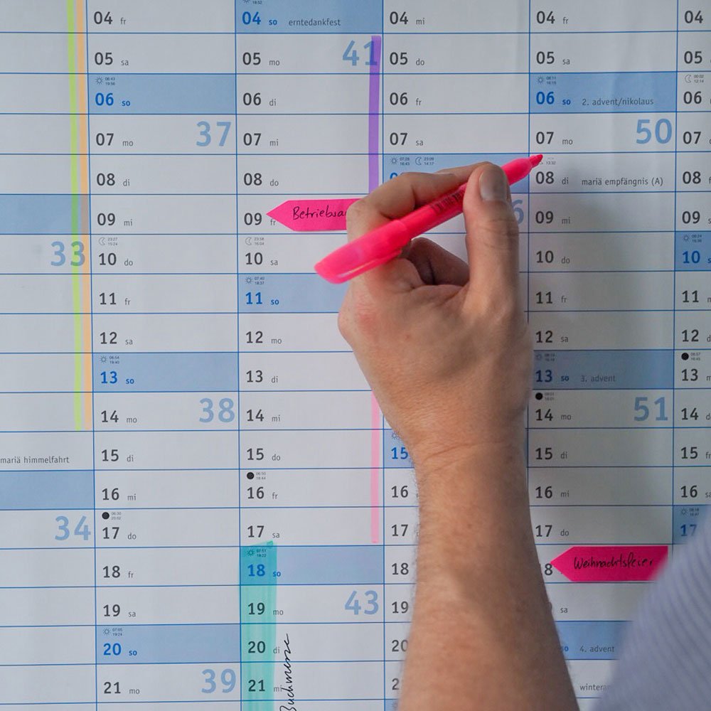 Eine Hand hält einen pinkfarbenen Marker. Dahinter ist ein Wandkalender zu sehen, auf dem mit Markern und Haftnotizen bereits einige Termine und Zeiträume gekennzeichnet wurden. 