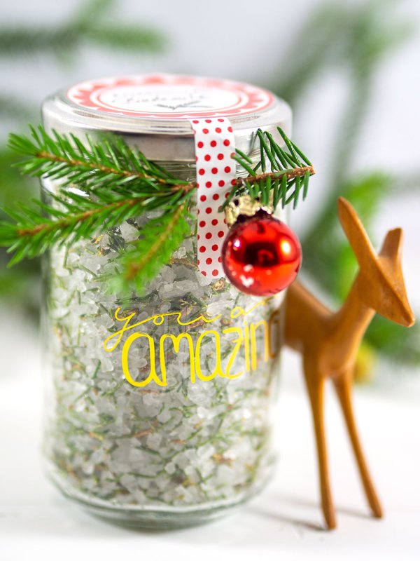 In einem Glas mit Schraubverschluss ist selbstgemachtes Fichtennadel-Badesalz eingefüllt. Das Glas ist schön beschriftet und mit einem kleinen Tannenzweig und einer kleinen Christbaumkugel dekoriert. Daneben steht ein kleines Reh aus Holz zur Deko.