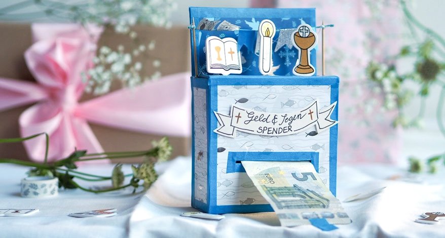 Auf einem mit Blumen geschmückten Tisch steht ein selbstgebasteltes Geschenk zu Kommunion bzw. Konfimation. Darauf sind verschiedene christiliche Symbole zu sehen und "Geld & Segenspender zu lesen". Ein 5€-Schein hängt heraus. Im Hintergrund sind verschwommen ein Geschenk und verschiedene weiße Blumen zu sehen.