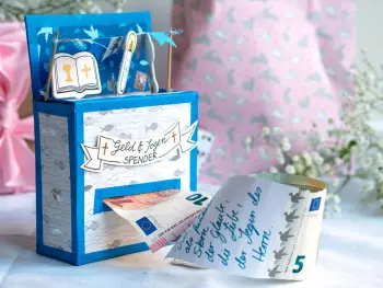 Ein selbstgebastelter "Geld und Segen Spender" aus Karton ist zu sehen. Er wurde mit Motivkarton mit Fisch-Motiv, Streudeko mit christlichen Motiven und Dekoband verziert. Aus einer Öffnung treten neben Geldscheinen noch beschriftete Zettel mit Segenswünschen hervor.