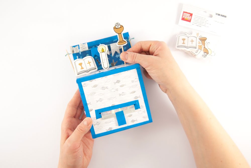 Auf einen mit Motivkarton und blauen Klebeband dekorierten Pappschachtel wird mit Hilfe von Zahnstochern Streudeko von Knorr Prandell geklebt. Diese zeigt eine Bibel, eine Kerze und einen Kelch. Im Hintergrund ist eine geschlossene Packung mit Streudeko zu sehen.