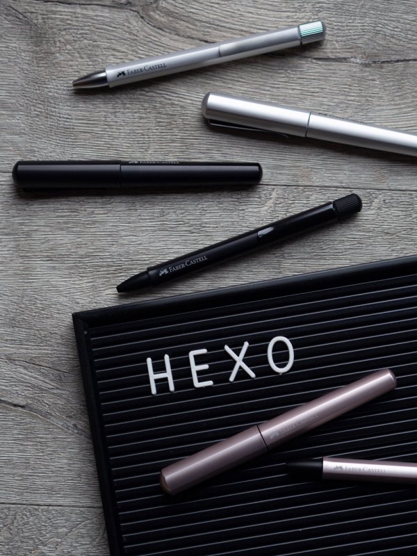 Auf einem grauen Holzboden liegt eine Buchstabentafel, auf der das Wort "HEXO" zu lesen ist. Im Bild verstreut liegen Kugelschreiber und Füller der Marke Faber-Castell. Sie gehören zur Hexo-Reihe und sind drei verschiedenen Farben zu sehen. In schwarz, silber und rosé.