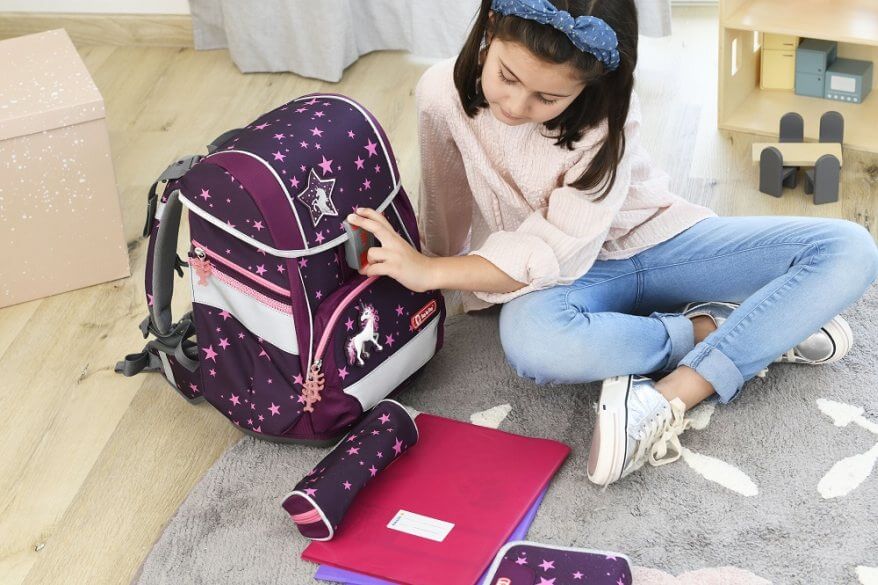 Ein Mädchen sitzt auf dem Boden ihres Kinderzimmers und hat ihre Schulsachen aus ihrem lila Step by Step Schulranzen ausgepackt, den sie gerade schließt. Vor ihr liegen ein Schulheft, ein Federmäppchen, ein Schlampermäppchen.