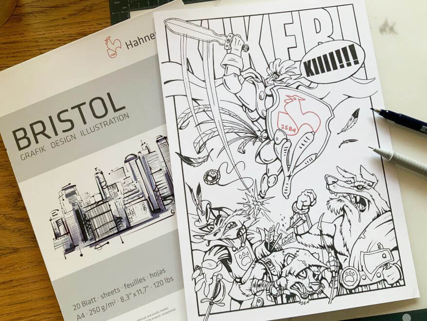 Auf einem Schreibtisch liegt die Reinzeichnung eines Comics in schwarz weiß. Das Motiv: Ein Hahn in Rüstung springt über Gegner hinweg. Im Hitnergrund liegt ein Block Bristolpapier von Hahnemühle.