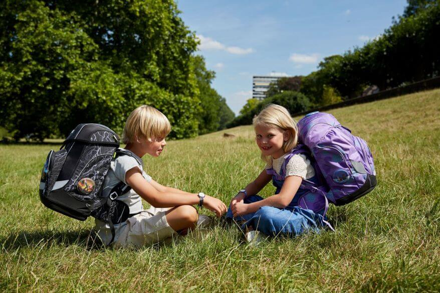Zwei Kinder sitzen im schneidersitz auf einer Wiese und unterhalten sich. Auf den Rücken tragen sie Schulranzen von ergobag