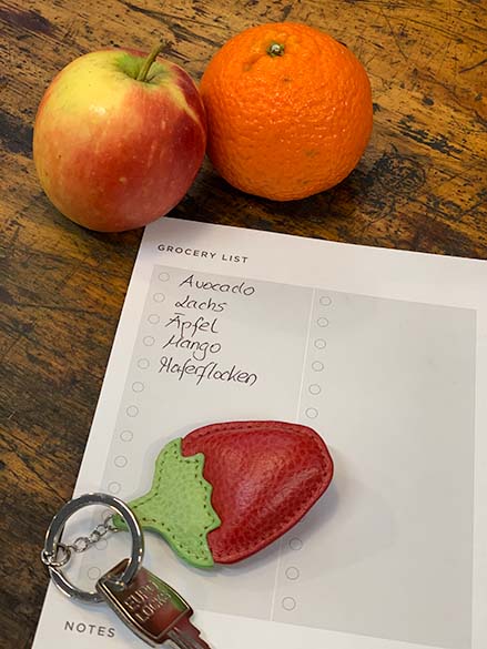 Auf einer Vorlage einer Einkaufsliste der Marke Filofax sind einige Produkte aufgelistet. Auf dem Notizpapier liegt ein Schlüssel mit einem Schlüsselanhänger in Form einer Erdbeere. Im oberen Bildrand liegen ein Apfel und eine Mandarine.