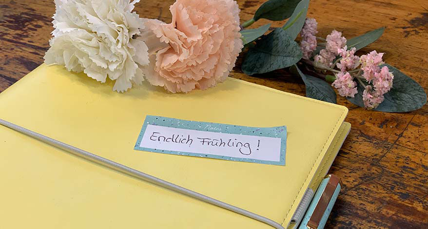 Auf einem Filofax-Kalender in pastellgelb klebt eine Haftnotiz mit den Worten „Endlich Frühling“. Am oberen Bildrand sieht man Blumen in rosa und weiß.