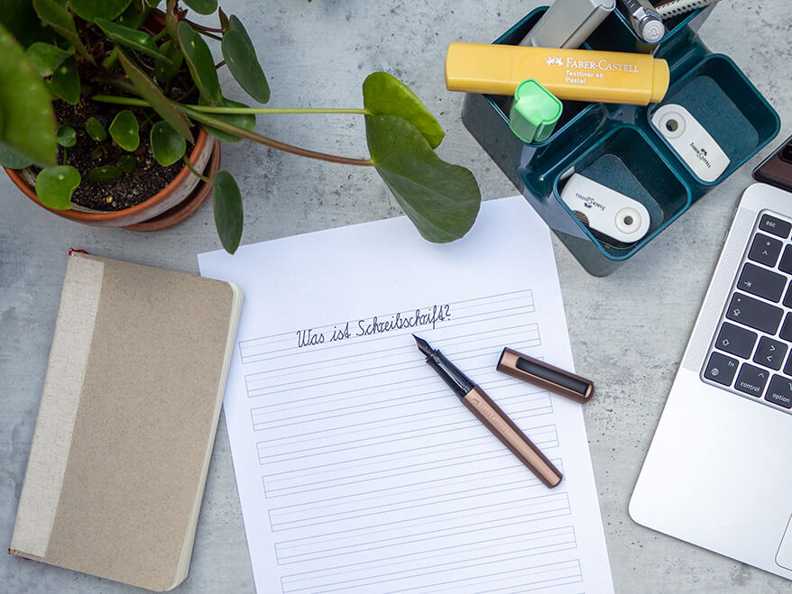 Auf einem Untergrund in Betonoptik sieht man eine Arbeitsplatz-Szenerie. In der Mitte liegt ein Blatt Papier mit der Aufschrift "Was ist Schreibschrift?", darauf ein Hexo-Füller der Marke Faber-Castell. Rechts im Bild ist ein Laptop zu sehen. Oberhalb des Blattes steht ein Stiftehalter, der mit diversen Materialien gefüllt ist. Links oben sieht man eine Zimmerpflanze ins Bild ragen, links unten liegt ein beiges Notizbuch. Foto: Johanna Rundel