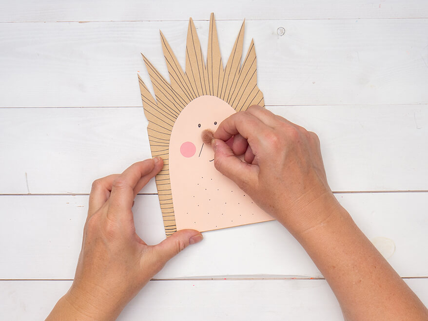 Jemand klebt einen braunen Mini-Pompon von Artoz als Igelschnauze auf eine zu einem Igel umgestaltete, braune Papiertüte. Der Untergrund ist weiß. Foto: Johanna Rundel