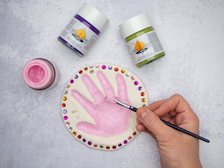 Jemand malt den Handabdruck aus Salzteig mit rosa Acrylfarbe und einem Pinsel an. Um den Handabdruck sind Glitzersteine von Knorr Prandell geklebt. Der Untergrund ist Betongrau. Foto: Johanna Rundel
