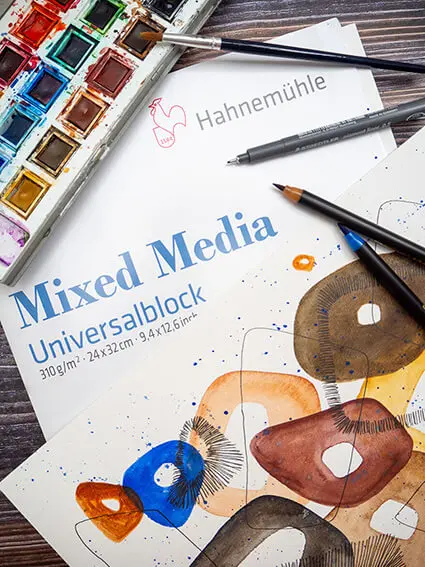 Auf einem dunklen Holztisch liegt ein Mixed-Media-Aquarellblock von Hahnemühle sowie ein fertiges Aquarellbild. Pinsel und Aquarellfarbkasten liegen dabei. Foto: Johanna Rundel