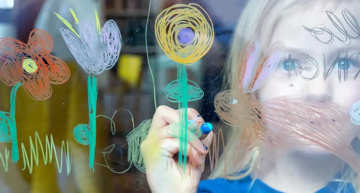 Ein Mädchen im Kindergartenalter steht am Fenster und malt mit einem woody-Buntstift von STABILO bunte Blumen auf die Scheibe. Foto: Johanna Rundel