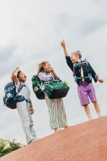 Drei Mädchen mit Schulranzen von ergobag stehen auf einem Hügel.