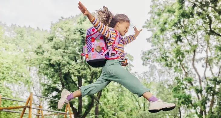 Ein Mädchen mit einem Schulranzen von ergobag auf dem Rücken springt mit weit ausgebreiteten Armen.
