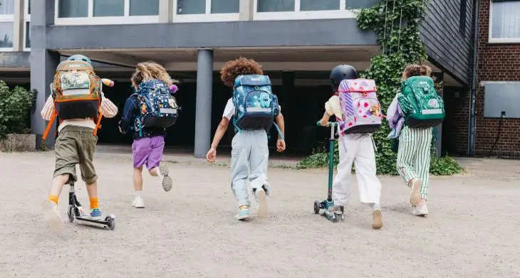 Rückenansicht von 5 Kindern, die mit Schulranzen von ergobag zu einer Schule laufen.