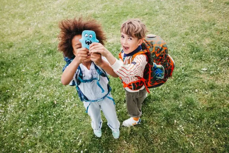 Zwei Kindern mit Schulranzen von ergobag stehen auf einer Wiese und halten ein Flausch-Klettie nach oben.