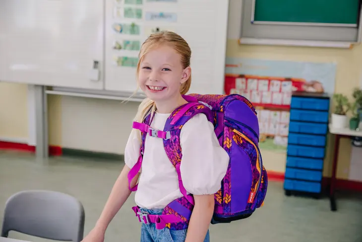 Ein Mädchen steht in einem Klassenzimmer und blickt lächelnd in die Kamera. Sie hat einen Schulranzen von Scout auf dem Rücken.