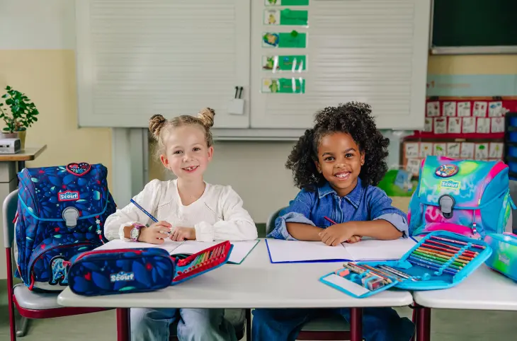 Zwei Mädchen sitzen an einem Schülertisch in einem Klassenzimmer. Neben ihnen stehen Schulranzen von Scout.