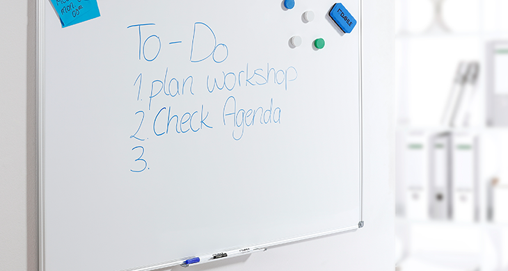 Das Bild zeigt ein Dahle-Whiteboard mit einer handgeschriebenen To-Do-Liste, die Aufgaben wie „Plan Workshop“ und „Check Agenda“ enthält. An der Tafel sind Magneten, ein Schwamm und Haftnotizen angebracht. Im Hintergrund sind Büroordner unscharf zu erkennen.