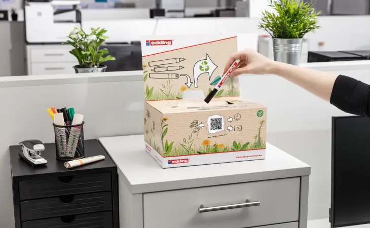 Eine Person hält einen schwarzen edding Marker über eine edding-Sammelbox aus Pappe mit Schlitz, die auf einem Schreibtisch steht. Die Box ist mit Zeichnungen zum Thema Umwelt versehen. Im Hintergrund sind Büromaterialien zu sehen.