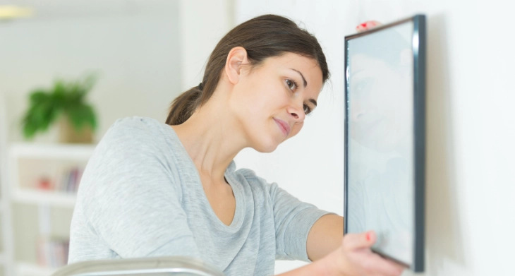 Frau in grauem Shirt hängt ein Bild mit schwarzem Rahmen an eine weiße Wand. Sie steht auf einer Leiter und richtet das Bild sorgfältig aus.