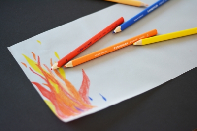 Auf einem Blatt Papier sind mit gelben, roten und orangen Buntestiften Flammen gemalt. Die Buntstifte der Marke Staedtler liegen auf dem Papier.