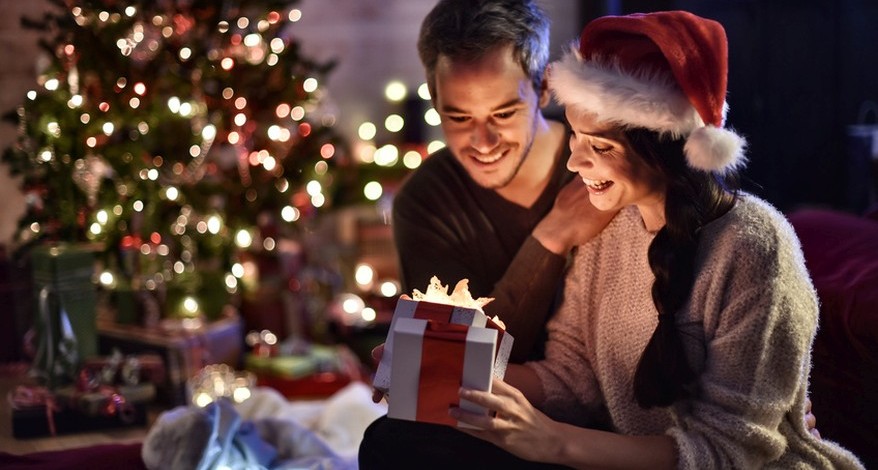 Ein junges Paar sitzt vor einem geschnücktem und beleuchteten Weihnachtsbaum. Die Frau hat eine Nikolausmütze auf dem Kopf und packt eine Geschenkschachtel aus. Beide schauen auf die Schatel und lächeln.