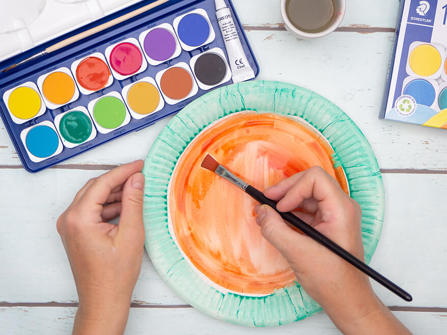 Jemand malt einen Pappteller mit Wasserfarben von Staedtler an. Innen wird der Teller rot angemalt, der Rand grün – wie eine Wassermelone. Der Untergrund ist hell. Foto: Johanna Rundel 