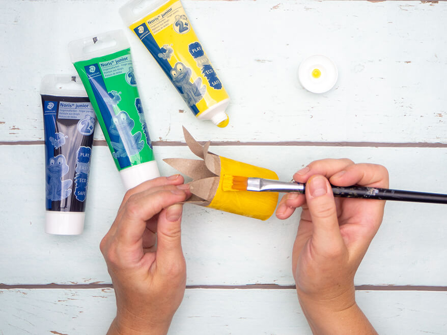 Jemand malt mit Fingerfarben von Staedtler eine Klopapierrolle zu zwei Drittel gelb an. Das obere Drittel der Rolle hat Zacken. Farbtuben liegen verteilt herum. Der Untergrund ist hell. Foto: Johanna Rundel 