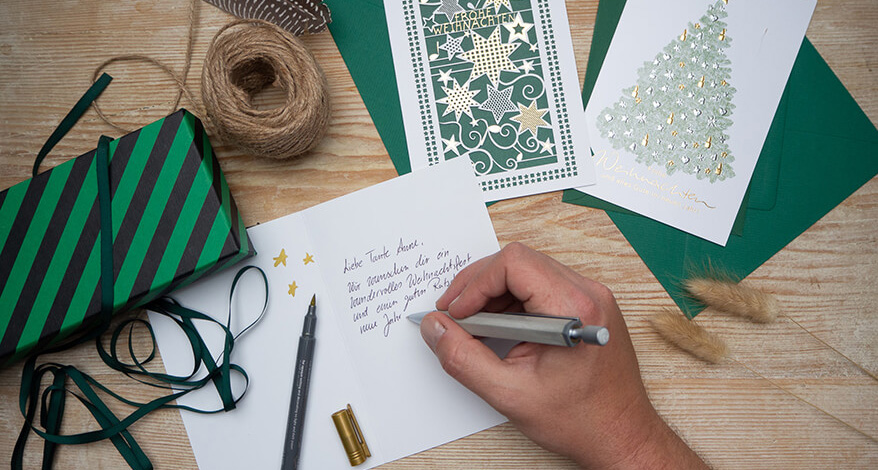 Auf einem Holztisch liegen verschiedene Weihnachtkarten von ABC Grußkarten, sowie grüne Briefumschläge. Die Karten sind mit aufwändiger Goldprägung in unterschiedlichen Designs gestaltet. Jemand schreibt mit einem Kugelschreiber Weihnachtsgrüße auf die Innenseite einer Karte. Ein geöffneter goldener Brushpen liegt daneben. Außerdem sind eine grün-schwarz gestreifte Geschenkschachtel sowie Geschenk- und Juteband zu sehen. Foto: Johanna Rundel