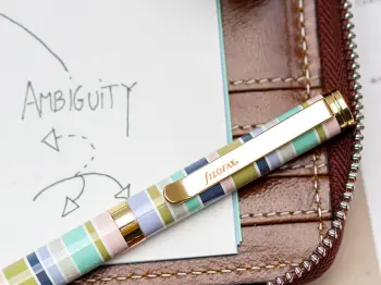 Auf einem braunen Filofax-Planer liegt ein gestreifter Kugelschreiber in Pastellfarben. Auf dem Einlegeblatt darunter steht: ambuguity. Foto: Johanna Rundel
