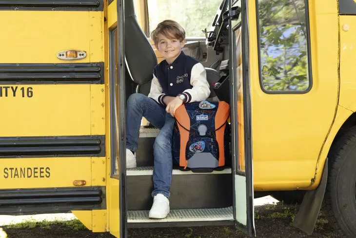Ein Kind sitzt in der geöffneten Tür eines gelb-schwarzen Schulbusses, neben ihm steht ein Schulranzen von Scout