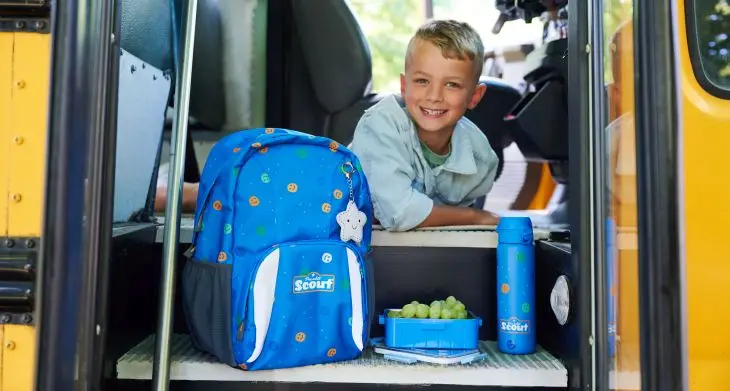 Ein Kind schaut aus der geöffneten Tür eines Schulbusses heraus, vor ihm steht ein Schulranzen, eine Brotdose und eine Trinkflasche von Scout